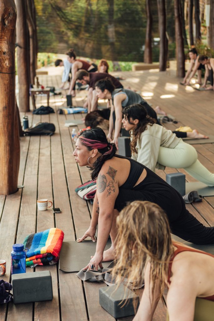 A group of students on a retreat in Mexico take a step forward while moving through sun salutations on a yoga deck overlooking the Pacific Ocean